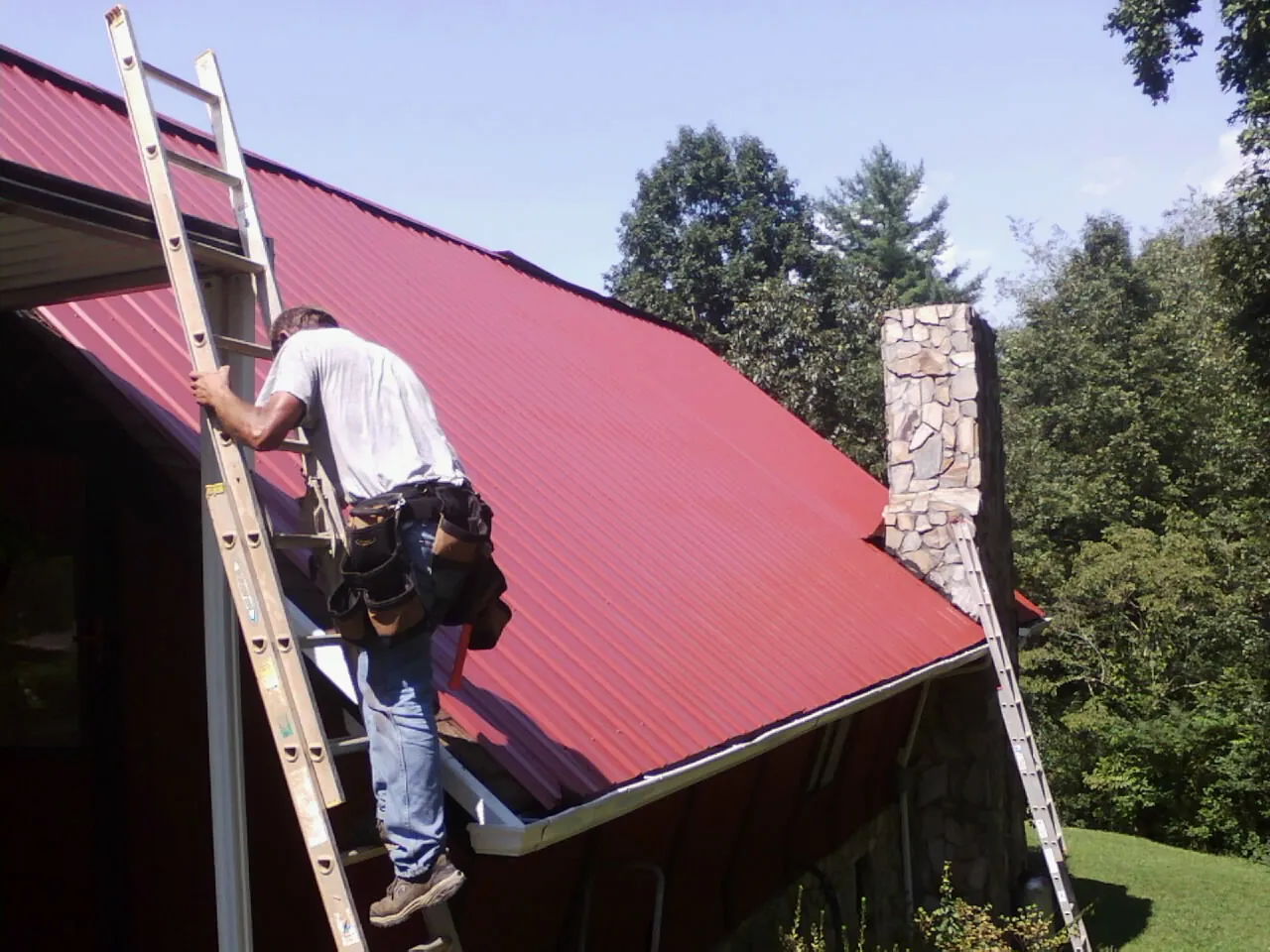 Roofer on a ladder beside a red metal roof and stone chimney — Soffit & Fascia Repair by Roofing Brothers in Crystal Bay